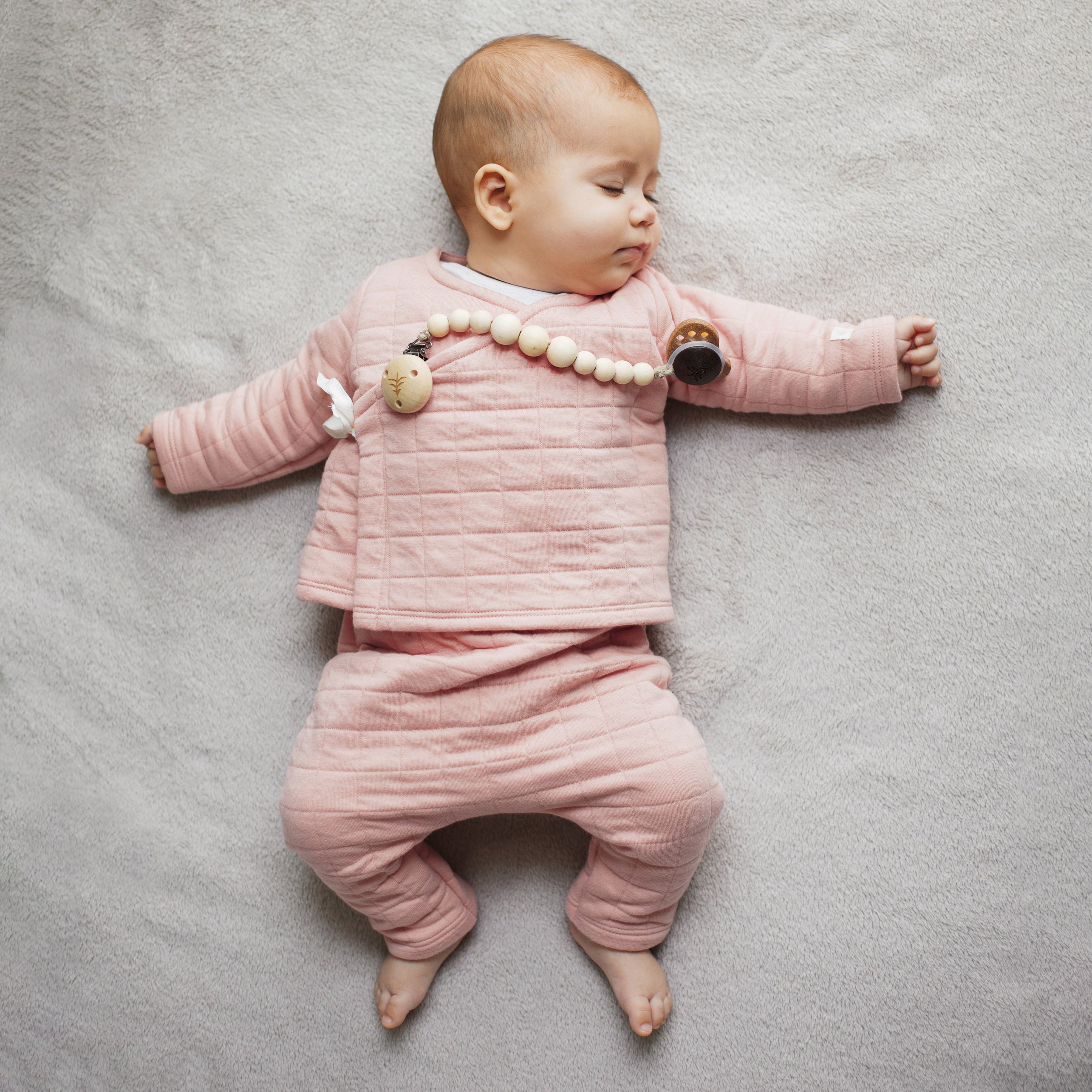 Sovande bebis klädd i mjuka rosa kläder med en naturlig napphållare i trä från Skogen Baby, praktisk och säker för små barn.